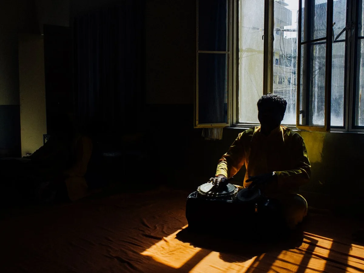 A musician seated on the floor playing tabla in a quiet room, warm afternoon light falling across the mat