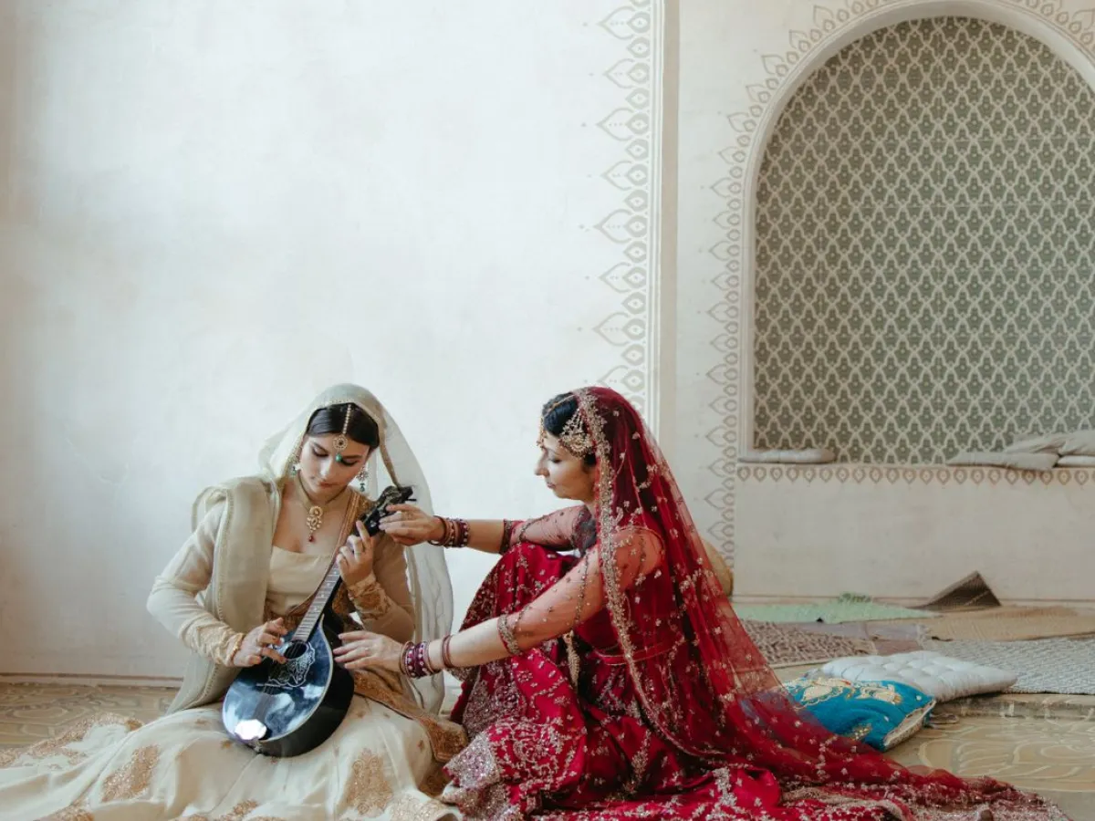 Two musicians in traditional dress seated on the floor of a warm, arched room, one tuning a stringed instrument